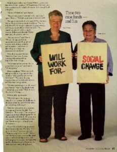 Harriet Miller and Monica Wehrle in a photo for the article in the Fort Wayne Magazine titled “The Dynamic Duos of 2005.” They are both smiling and holding up signs that say “will work for… social change.” Fort Wayne, Indiana, 2005. Photo Courtesy of Harriet Miller and Monica Wehrle.