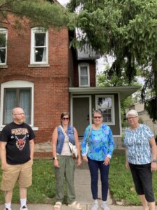 L-R: Justin, Rikki, Peggy, and Monica Wehrle in front of Monica’s childhood brick house. In Fort Wayne, Indiana, 2024. Photo Courtesy of Harriet Miller and Monica Wehrle.